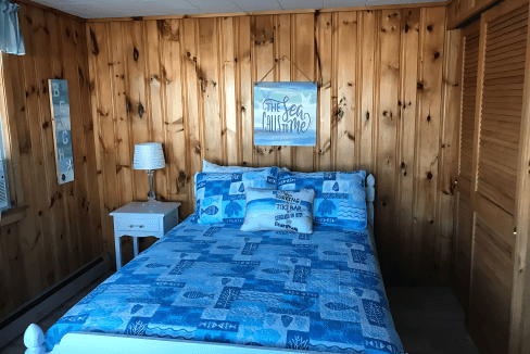 Bedroom with wood-paneled walls featuring a bed with blue patterned bedding, a side table with a lamp, and a wall art piece above the bed.