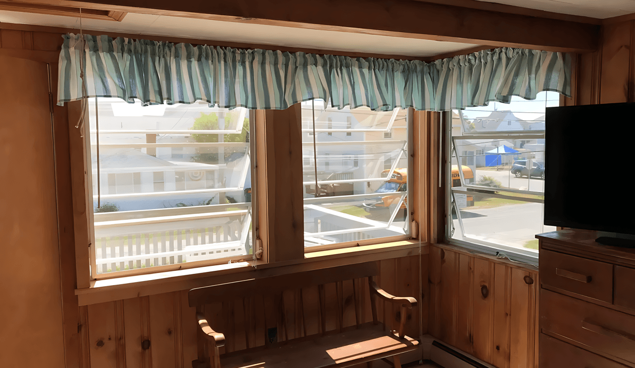 Room with two windows and wooden walls, featuring striped curtains. A wooden bench is placed beneath the windows and a TV is on the right. Outside, a glimpse of a street and houses is visible.