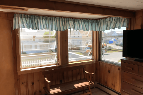 Room with two windows and wooden walls, featuring striped curtains. A wooden bench is placed beneath the windows and a TV is on the right. Outside, a glimpse of a street and houses is visible.