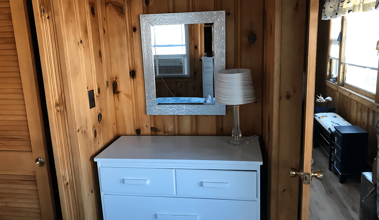 A white dresser with a mirror above it is positioned against a wooden-paneled wall. A lamp sits on the dresser. A partially open door reveals another room.