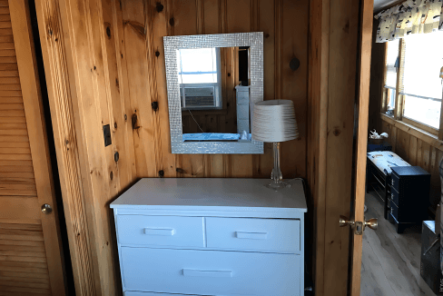 A white dresser with a mirror above it is positioned against a wooden-paneled wall. A lamp sits on the dresser. A partially open door reveals another room.