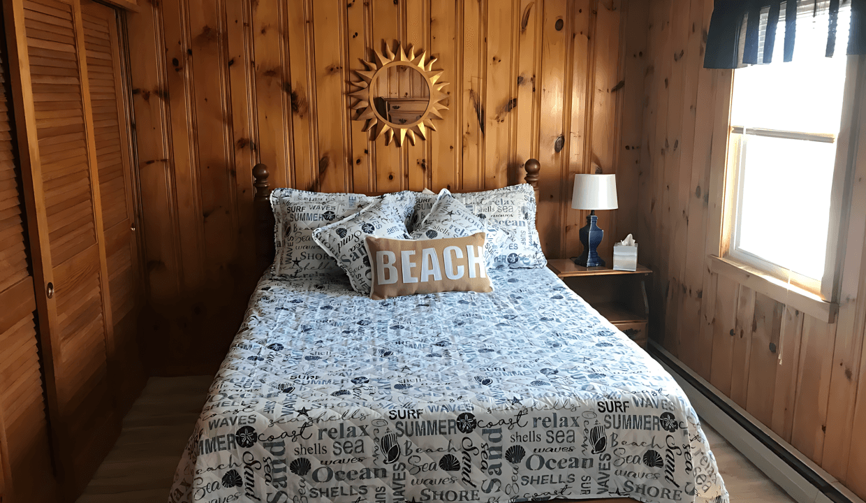 A bedroom with wood-paneled walls features a bed with beach-themed bedding, a round sunburst mirror, and a small wooden nightstand with a lamp. A window with a valance provides natural light.