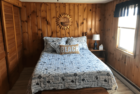 A bedroom with wood-paneled walls features a bed with beach-themed bedding, a round sunburst mirror, and a small wooden nightstand with a lamp. A window with a valance provides natural light.