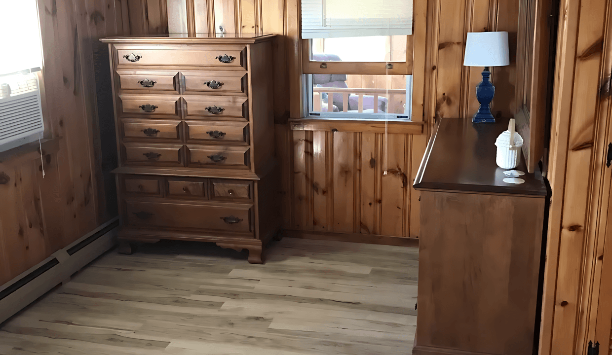 A room with wooden paneled walls, a ceiling fan, dressers, a window with a blue curtain, and light wood flooring.