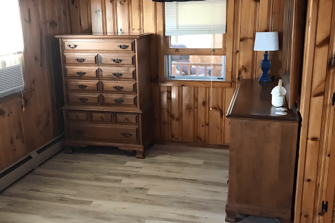 A room with wooden paneled walls, a ceiling fan, dressers, a window with a blue curtain, and light wood flooring.