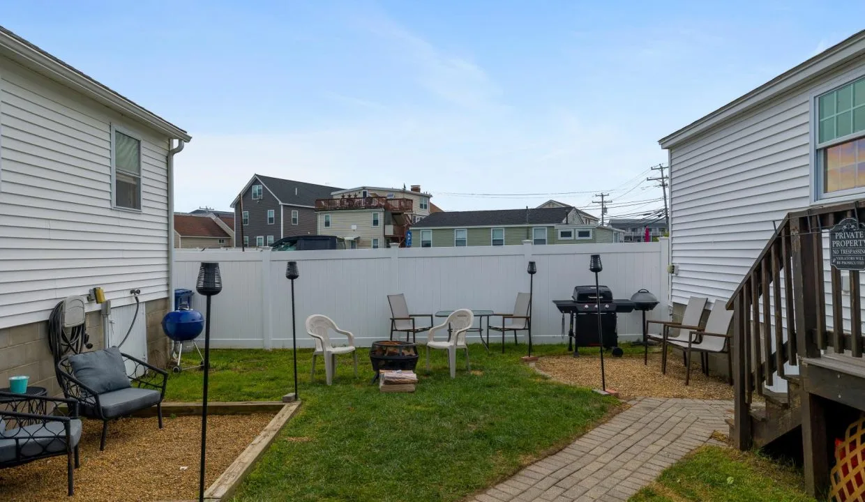 Backyard with chairs, a fire pit, grill, and some small structures, bordered by two white buildings and a white fence. There's a short path leading from a wooden deck.