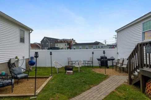 Backyard with chairs, a fire pit, grill, and some small structures, bordered by two white buildings and a white fence. There's a short path leading from a wooden deck.