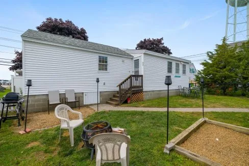 Single-story white house with a small porch, chairs, and a fire pit in the grassy yard. Water tower visible in the background.