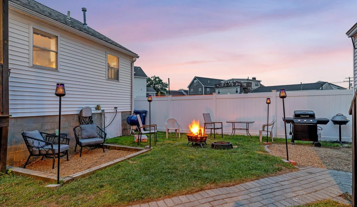 Backyard with a lit fire pit, surrounded by chairs, a grill, and tiki torches, set against a house with a white fence at sunset.