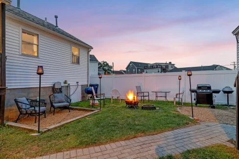 Backyard with a lit fire pit, surrounded by chairs, a grill, and tiki torches, set against a house with a white fence at sunset.