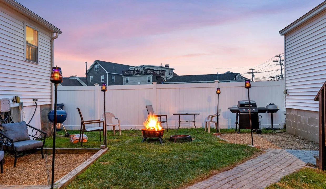 Backyard scene at dusk with a lit fire pit surrounded by chairs. Tiki torches are spaced around, and a barbecue grill is placed near one of the white houses flanking the yard.