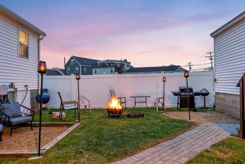 Backyard scene at dusk with a lit fire pit surrounded by chairs. Tiki torches are spaced around, and a barbecue grill is placed near one of the white houses flanking the yard.